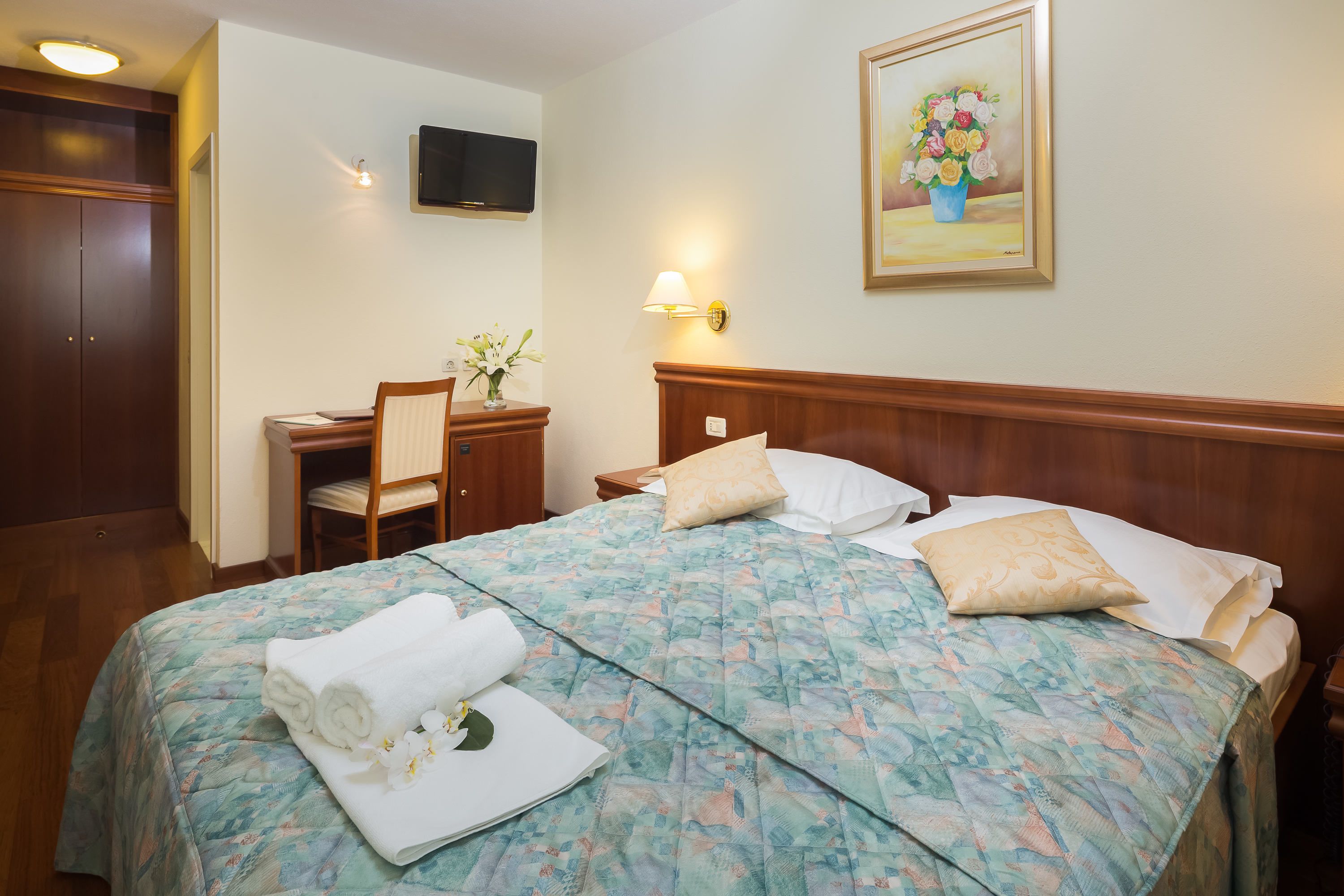 A hotel room with a bed, a desk, a chair, a television, and a painting on the wall.
