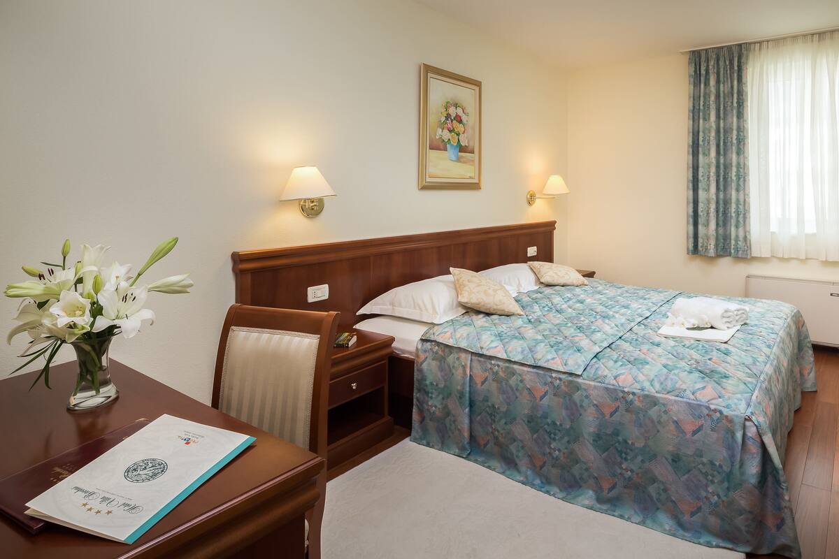 A hotel room with a wooden bed, two bedside lamps, a painting on the wall, a window with curtains, and a desk with a vase of flowers and a certificate.