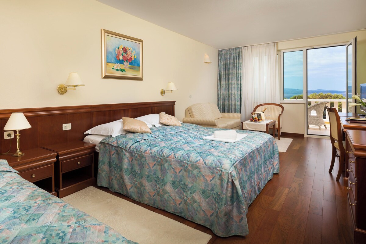 A hotel room with two beds, a sitting area with a chair, a table with a vase, and a painting on the wall.