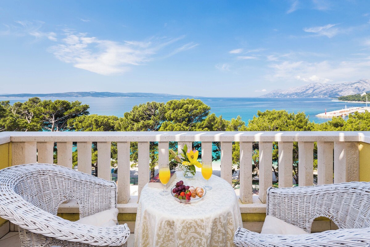 A balcony with wicker furniture, a table with fruit and drinks, and a scenic view of the sea and mountains.