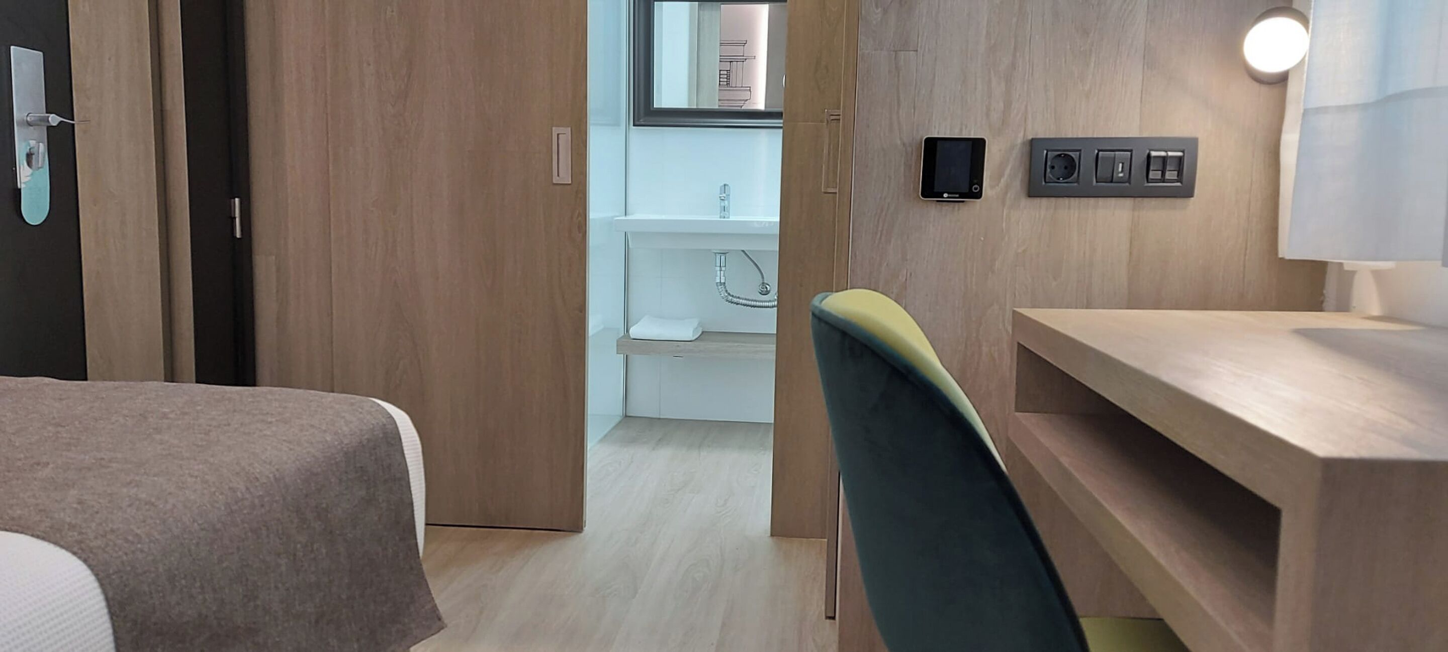 Une chambre d’hôtel avec une porte en bois, un bureau, une chaise, une table de chevet et un luminaire fixé au mur.
