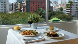 Een tafel met borden eten, een glas rode wijn en een kleine plant op een balkon met uitzicht op de stad.