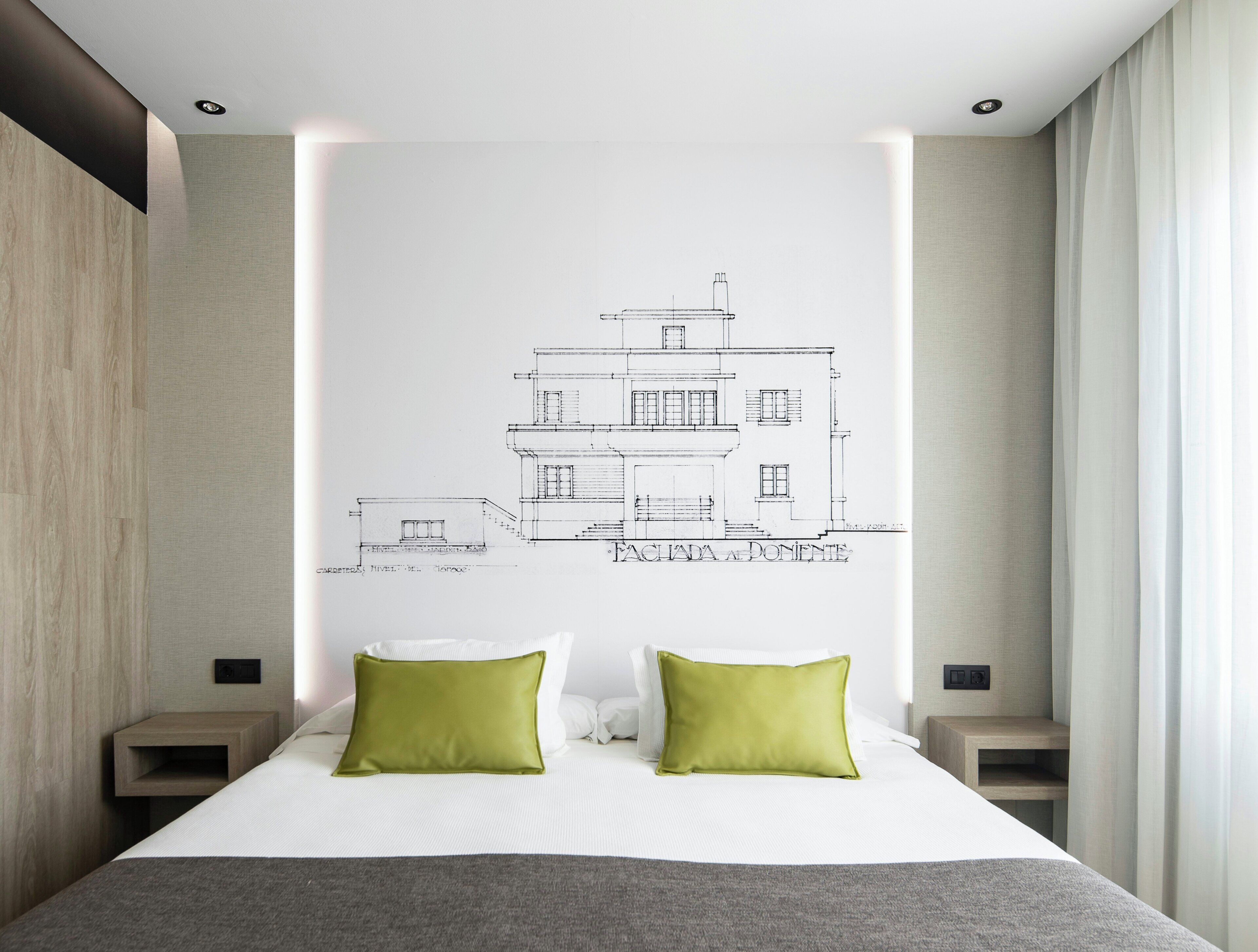 A bedroom with a bed, two green pillows, a white headboard, and a wall-mounted architectural drawing.