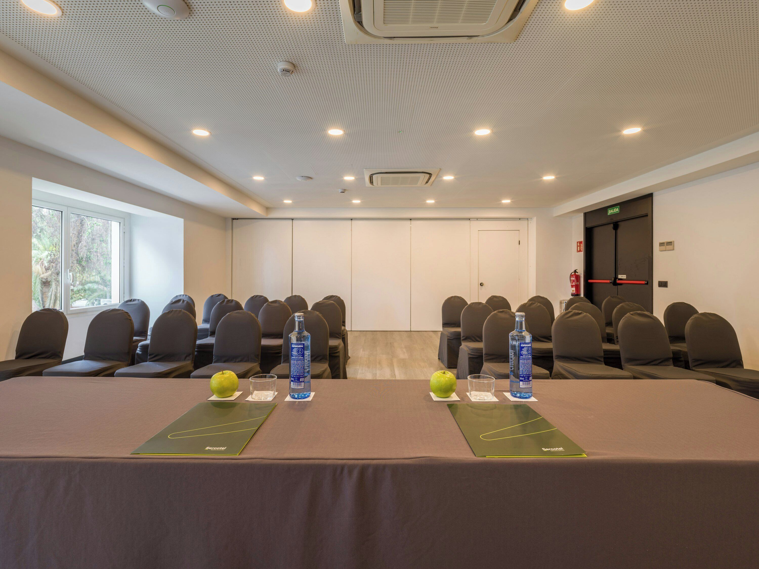 Een congreszaal met rijen stoelen, een tafel met waterflesjes en appels, en een deur met de aanduiding 'SALIDA'.
