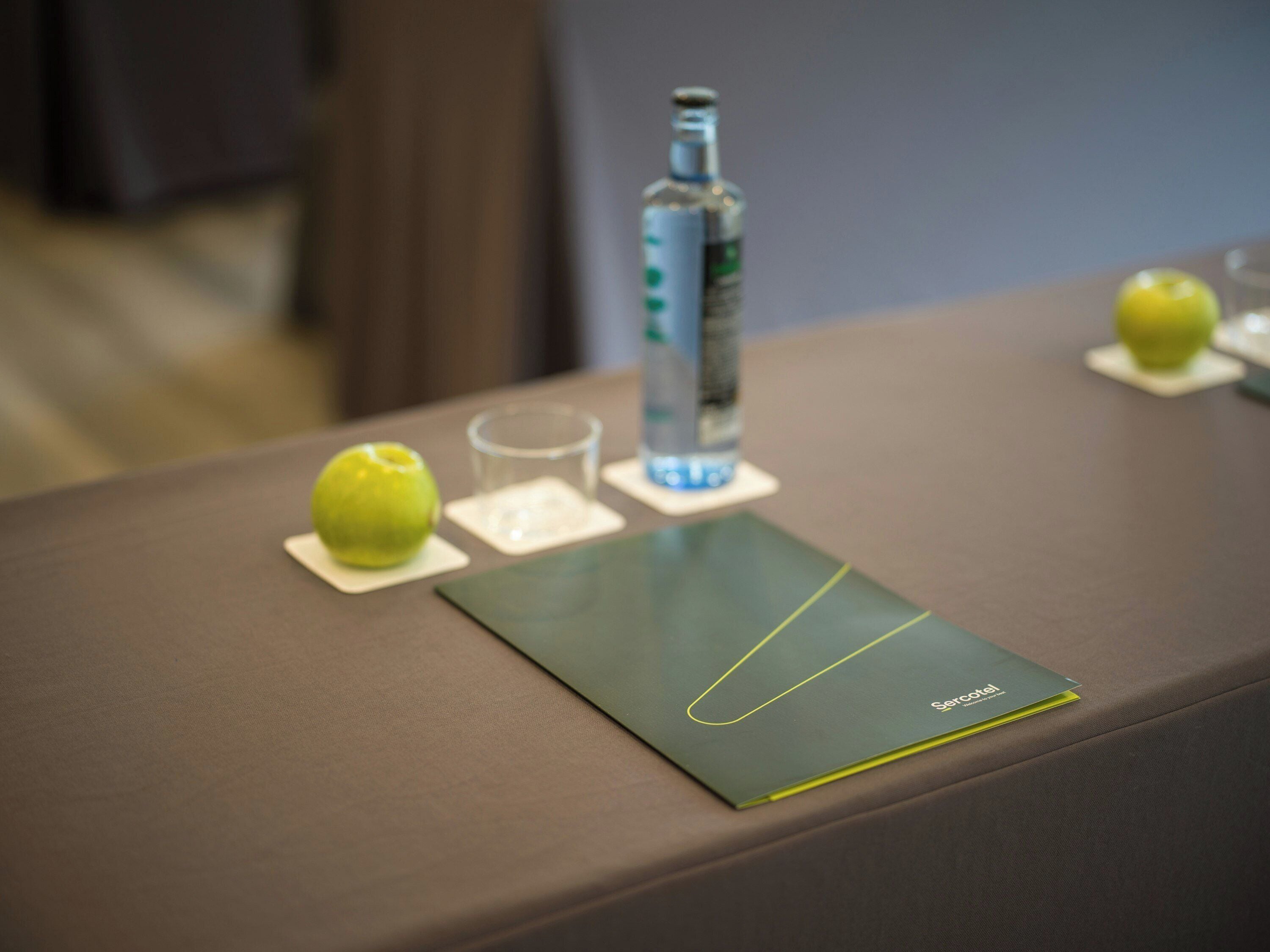 A table with a bottle, a glass, and an apple on coasters.