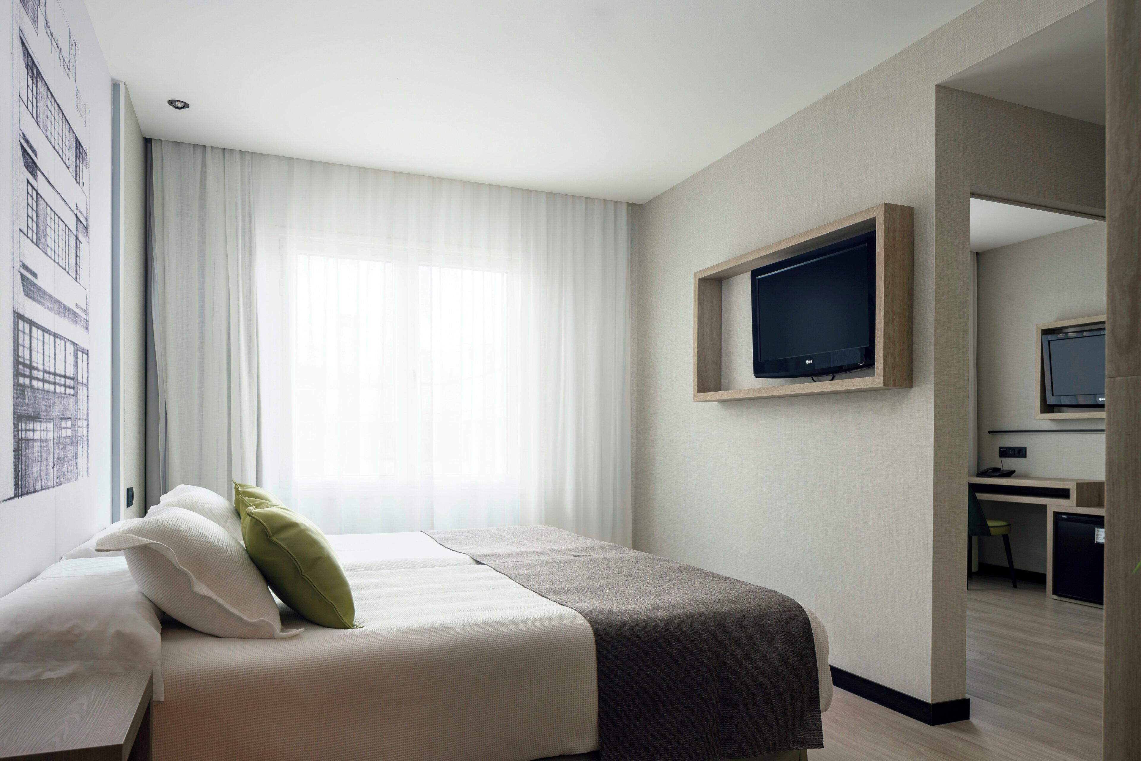 A hotel room with a bed, a TV mounted on the wall, a desk with a chair, and a window with curtains.