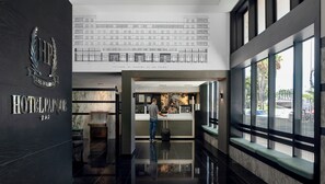 Un vestíbulo de hotel con mostrador de recepción, una escalera y un amplio ventanal con vistas a la calle.