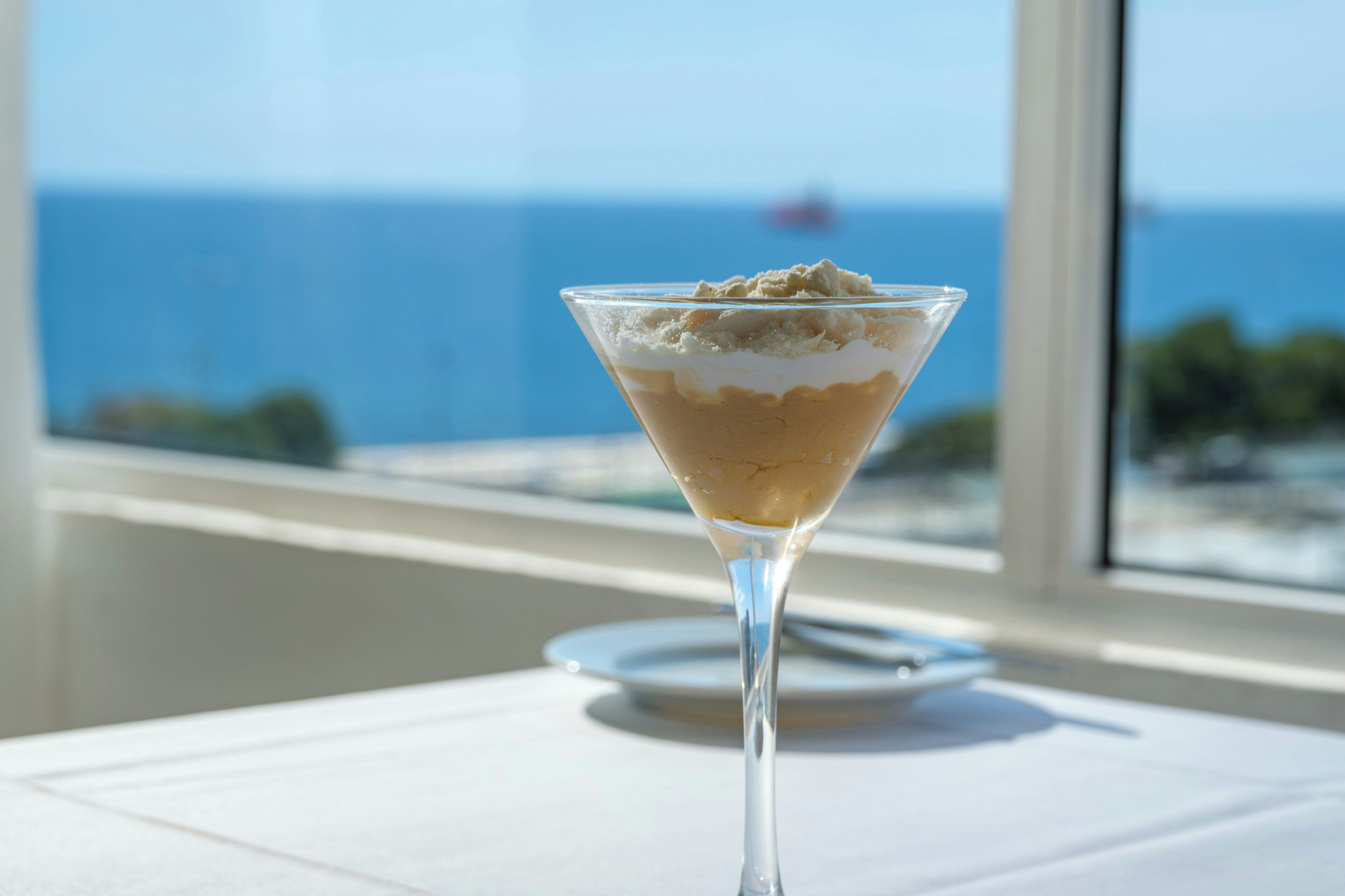 A martini glass with a dessert on a table by a window with a view of the ocean.