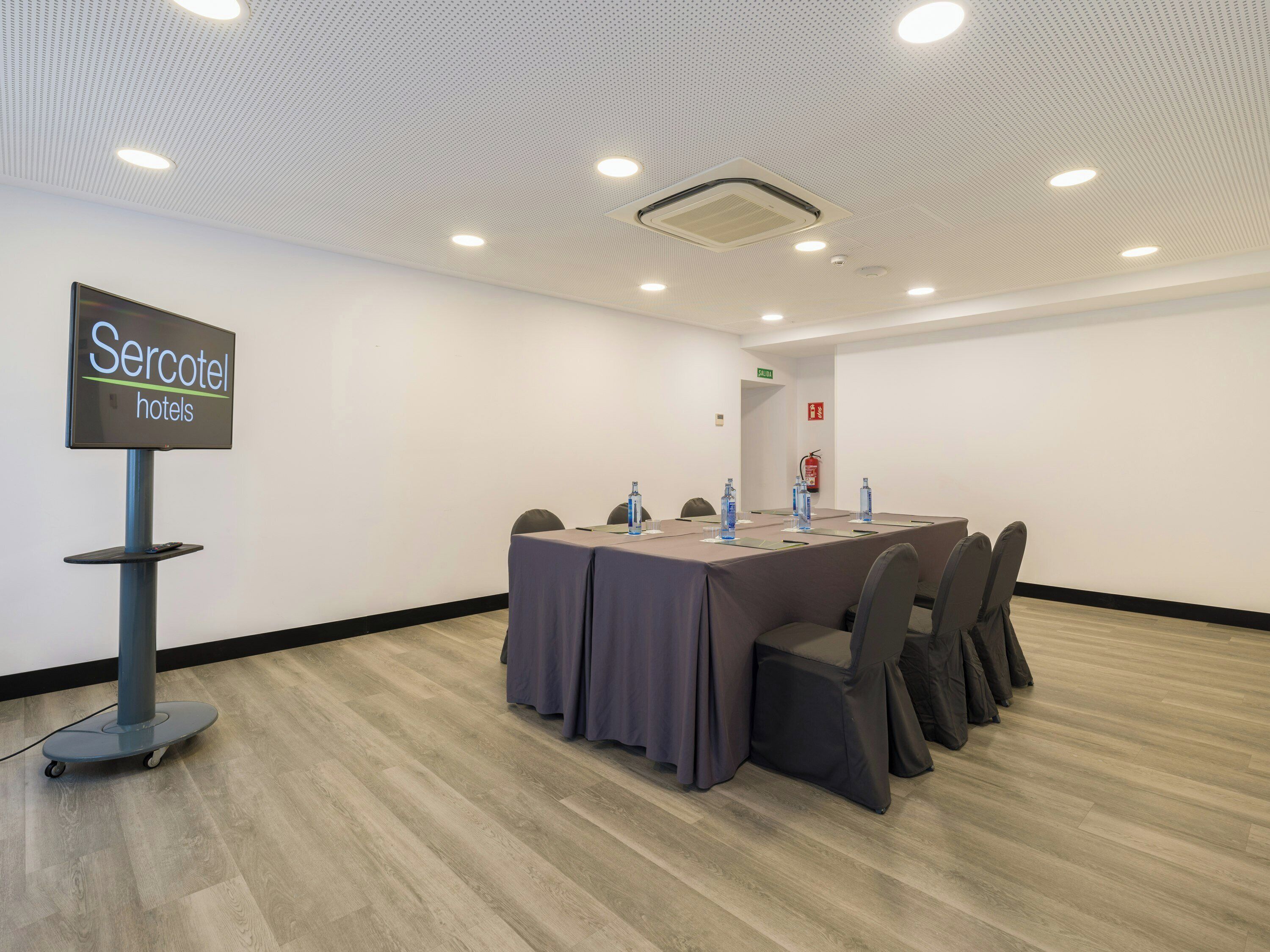 Una sala de conferencias con una larga mesa, sillas, un cartel de 'Sercotel hotels' y un extintor en la pared.