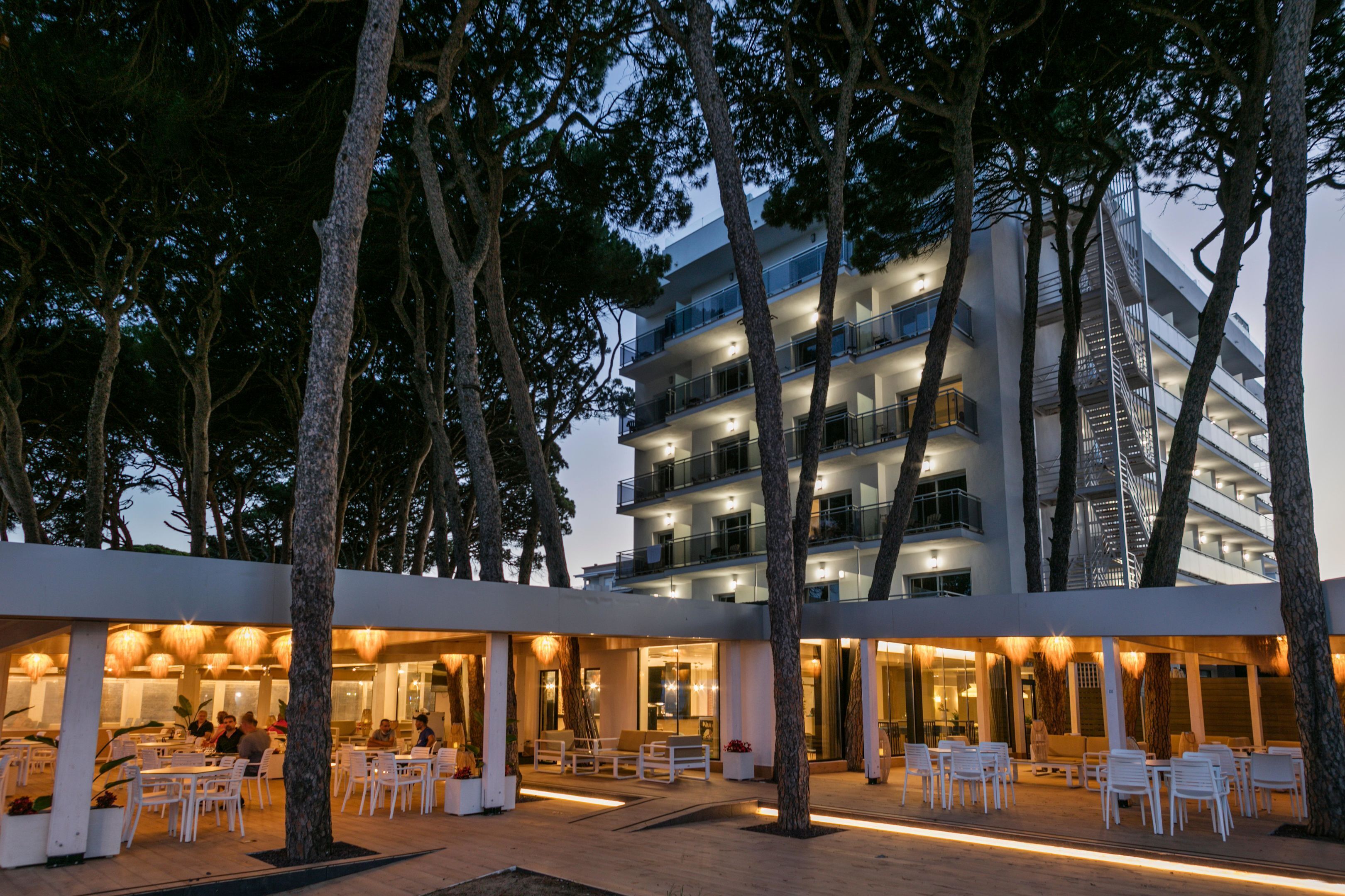 Een terras met witte stoelen en tafels, omringd door bomen, voor een modern gebouw met meerdere balkons.
