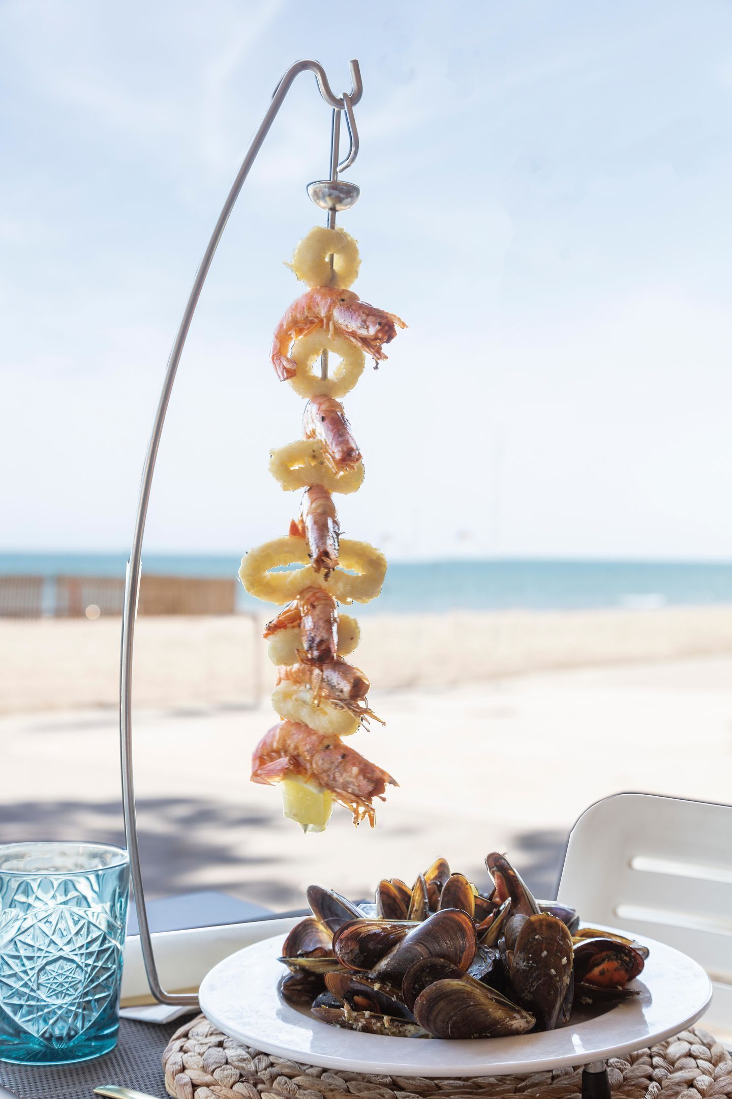 Een visgrill met garnalen en coquilles, een bord met mosselen, en een blauw glas op een strandtafel.