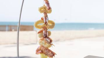 Een visgrill met garnalen en coquilles, een bord met mosselen, en een blauw glas op een strandtafel.