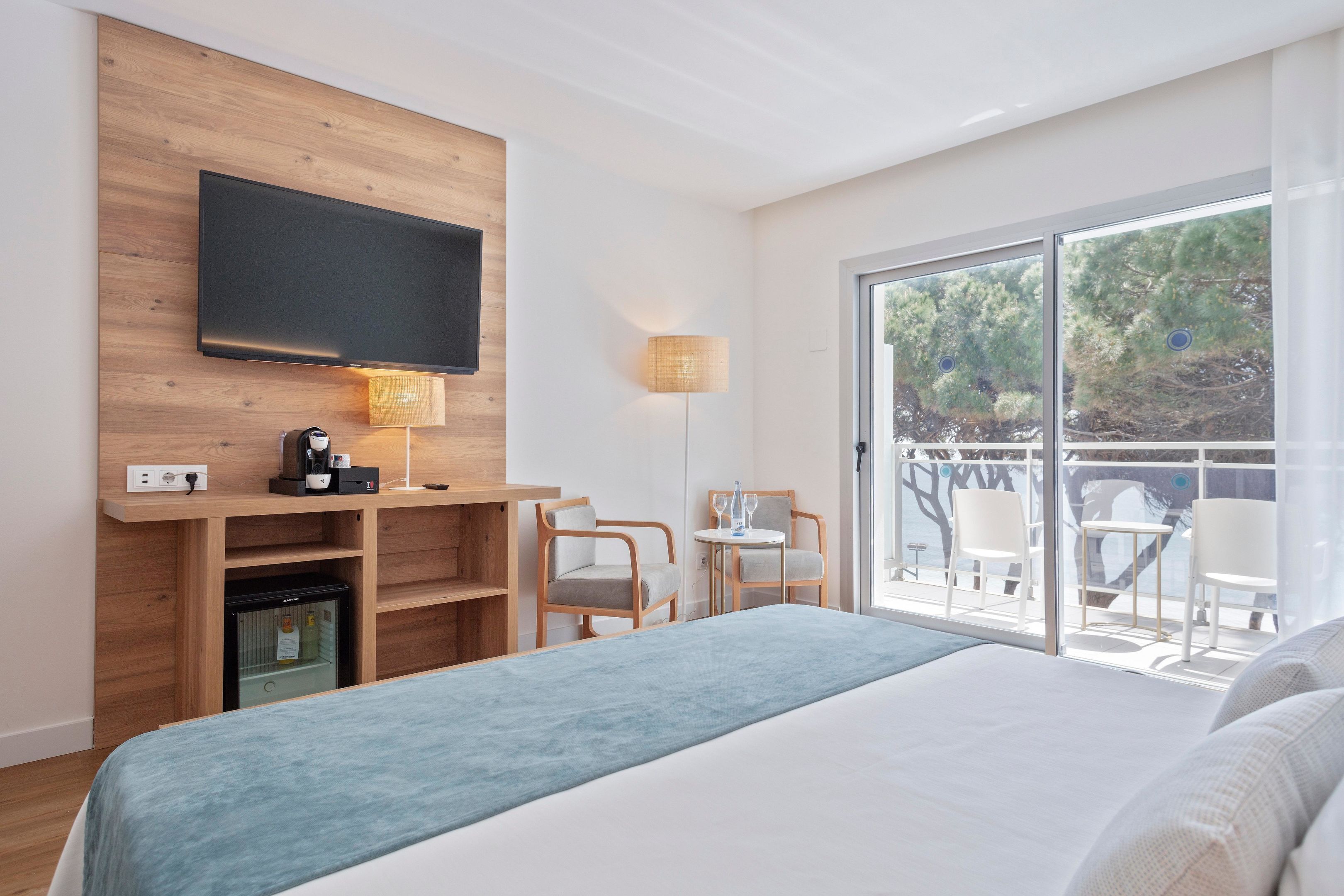 A modern hotel room with a large bed, a flat-screen TV mounted on a wooden panel, a small refrigerator, a desk with a coffee maker, and a balcony with outdoor seating.