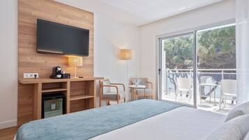 A modern hotel room with a large bed, a flat-screen TV mounted on a wooden panel, a small refrigerator, a desk with a coffee maker, and a balcony with outdoor seating.