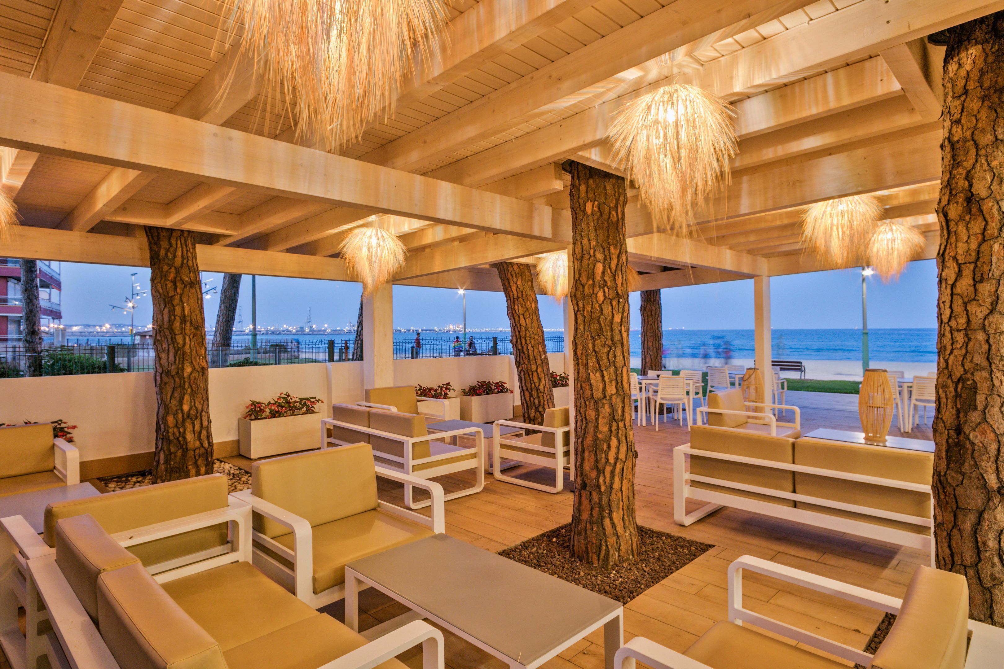 A beachside lounge with white furniture, large trees, and a view of the ocean.