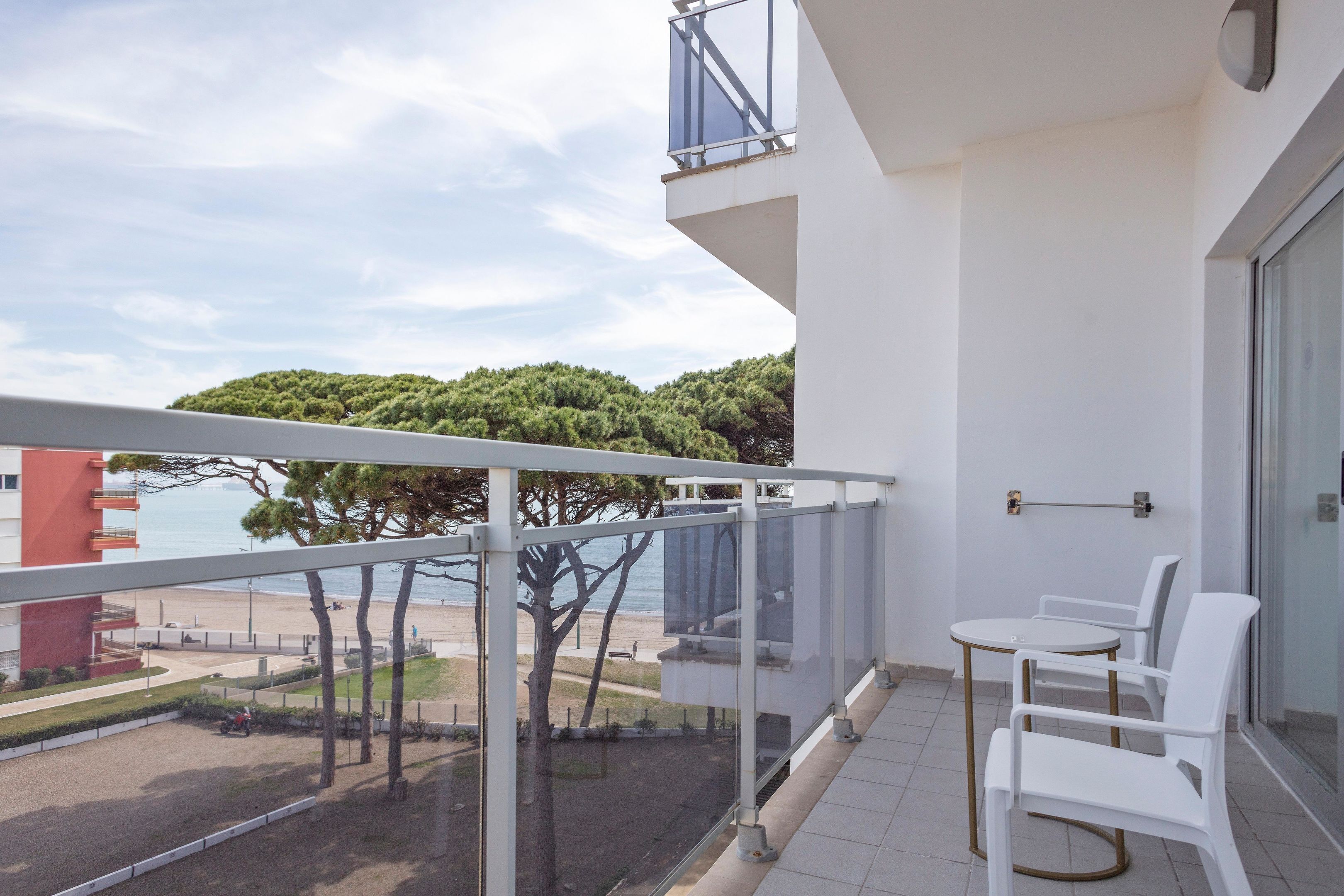 A balcony with a glass railing overlooking a beach and a forest. There are two white chairs and a small round table.
