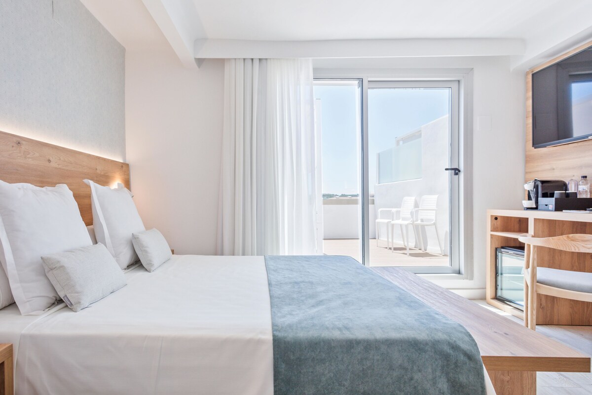 A modern hotel room with a large bed, a TV, and a balcony with outdoor seating.