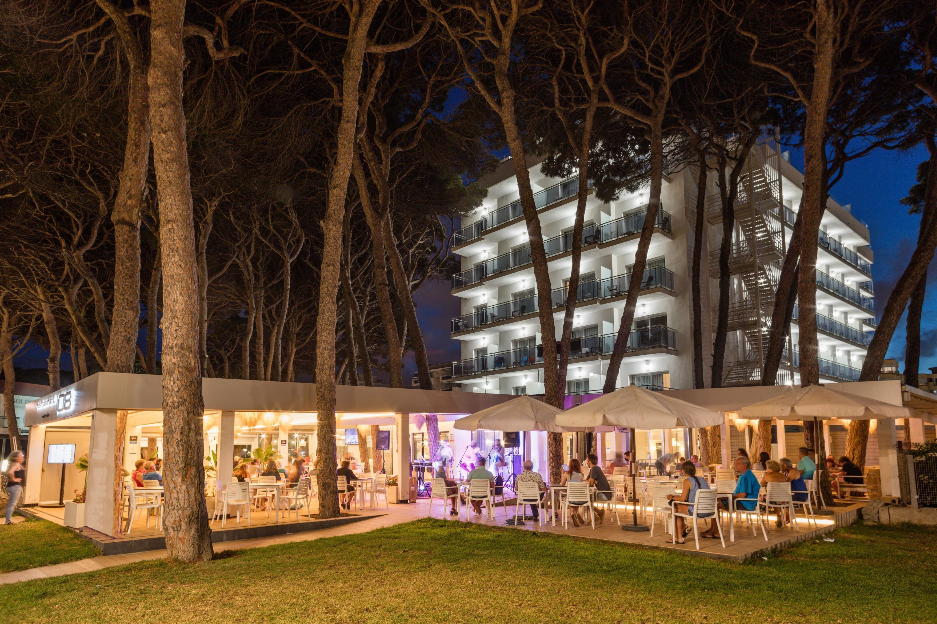 Un espace extérieur aménagé pour les repas, avec des tables et des chaises, entouré d’arbres et avec un bâtiment doté de plusieurs balcons en arrière-plan.