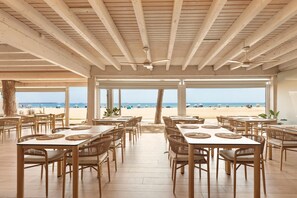A beachside restaurant with wooden tables and chairs, large windows, and ceiling fans. - Hotel Best Terramarina (Vila-Seca)