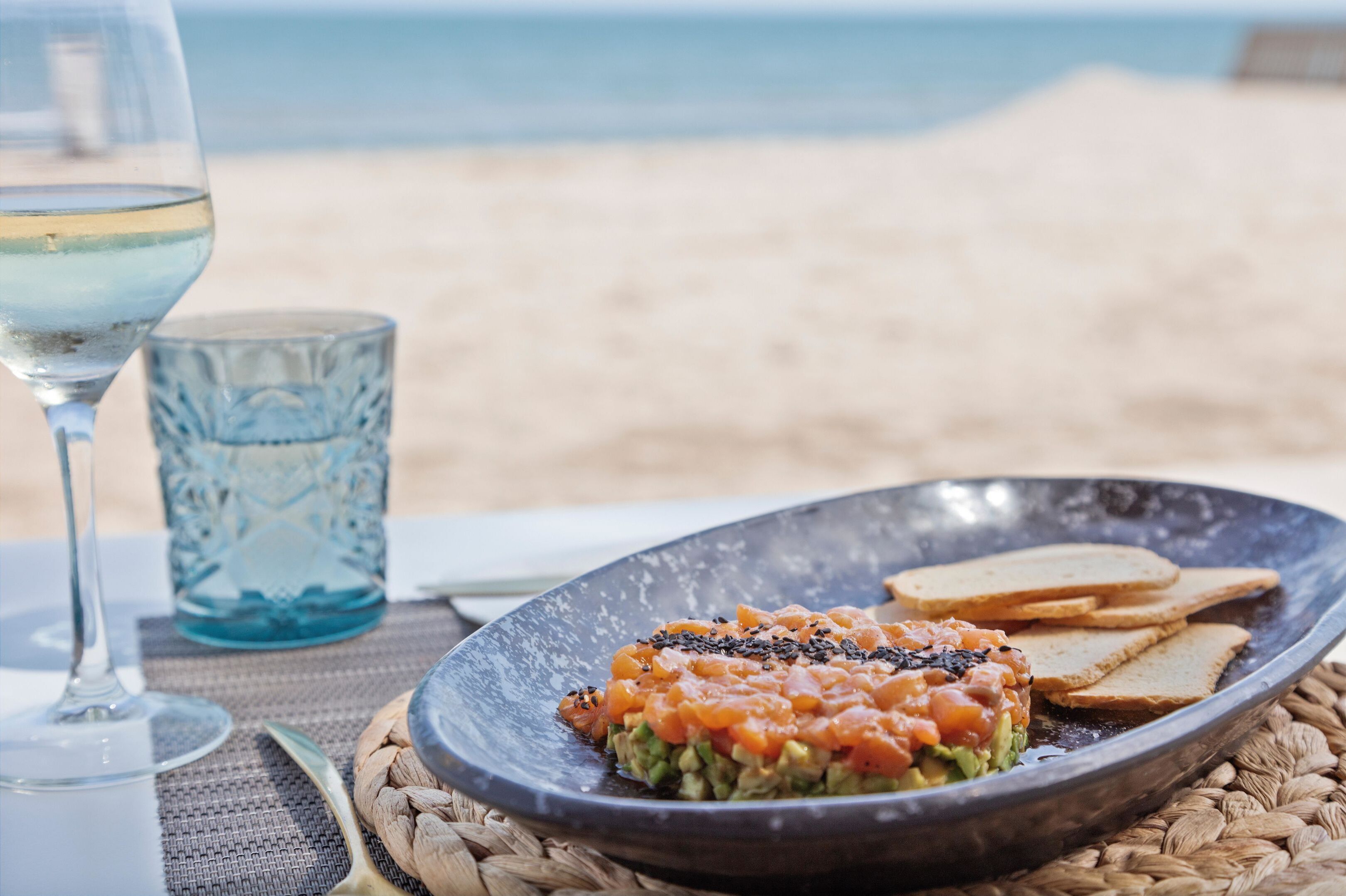 Een maaltijd op een geweven mat, met een glas witte wijn en een blauw glas ernaast, op het strand.