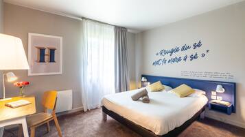 A hotel room with a bed, a desk with a lamp, a chair, and a wall with a quote.
