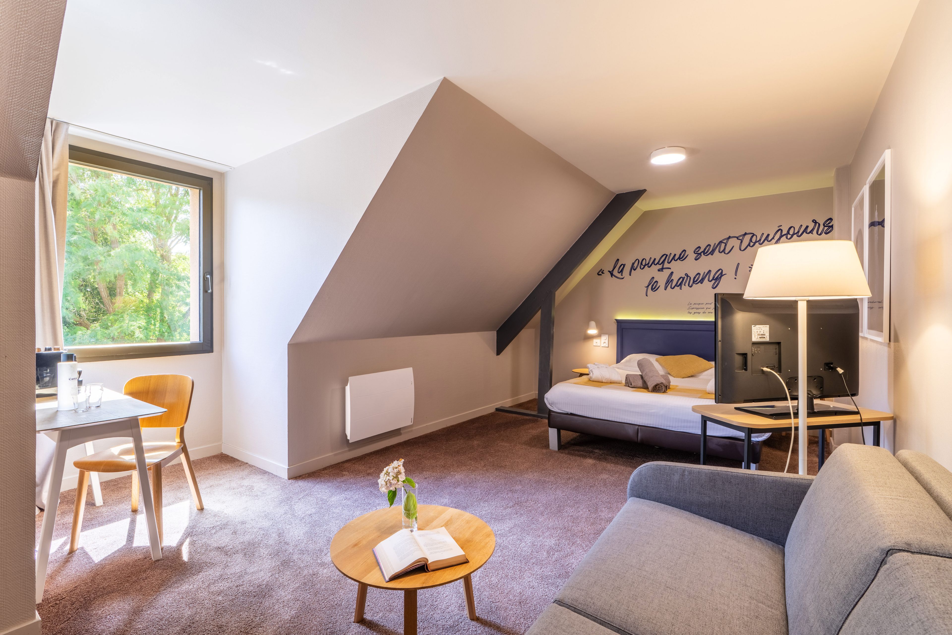 Un dormitorio moderno con techo inclinado, una cama, un escritorio, una silla, una mesita con un libro y una lámpara.
