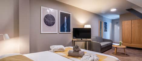Quarto de hotel com cama, televisão, sofá, guarda-roupa de madeira e duas imagens emolduradas na parede.