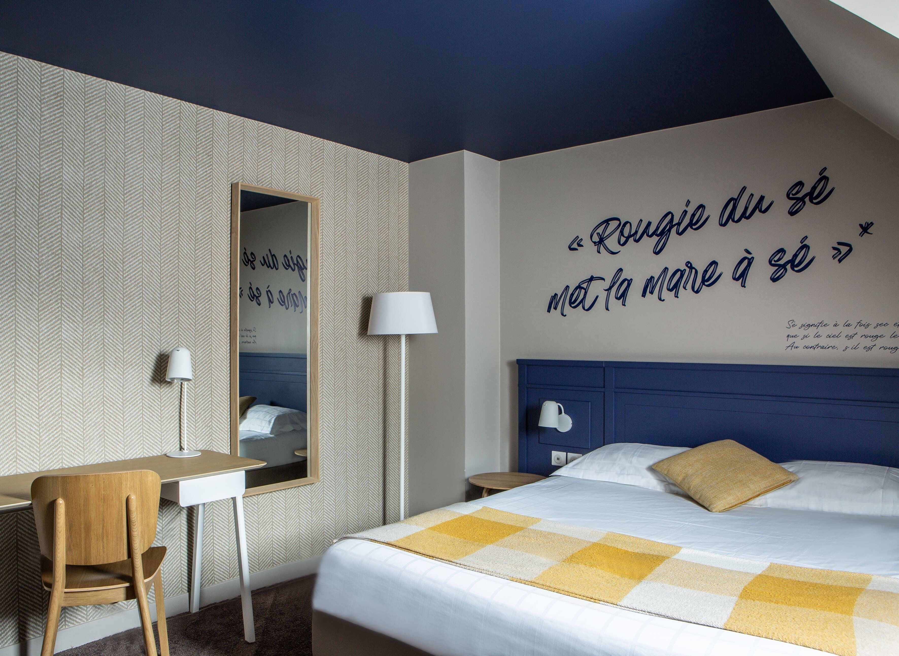 Ett hotellrum med en säng, ett skrivbord, en stol, en lampa och en vägg med ett citat.