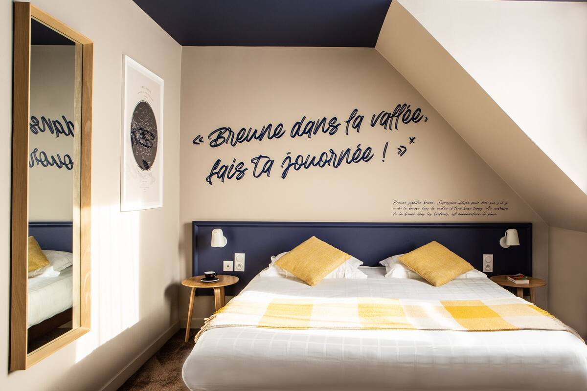 A bedroom with a bed, a mirror, a wall with a quote, and a small table with a cup.