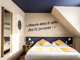 Quarto com cama, espelho, parede com uma frase e uma pequena mesa com uma xícara.