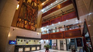 Un vestíbulo de hotel con una recepción, un mural grande, una escalera y una planta en el centro.