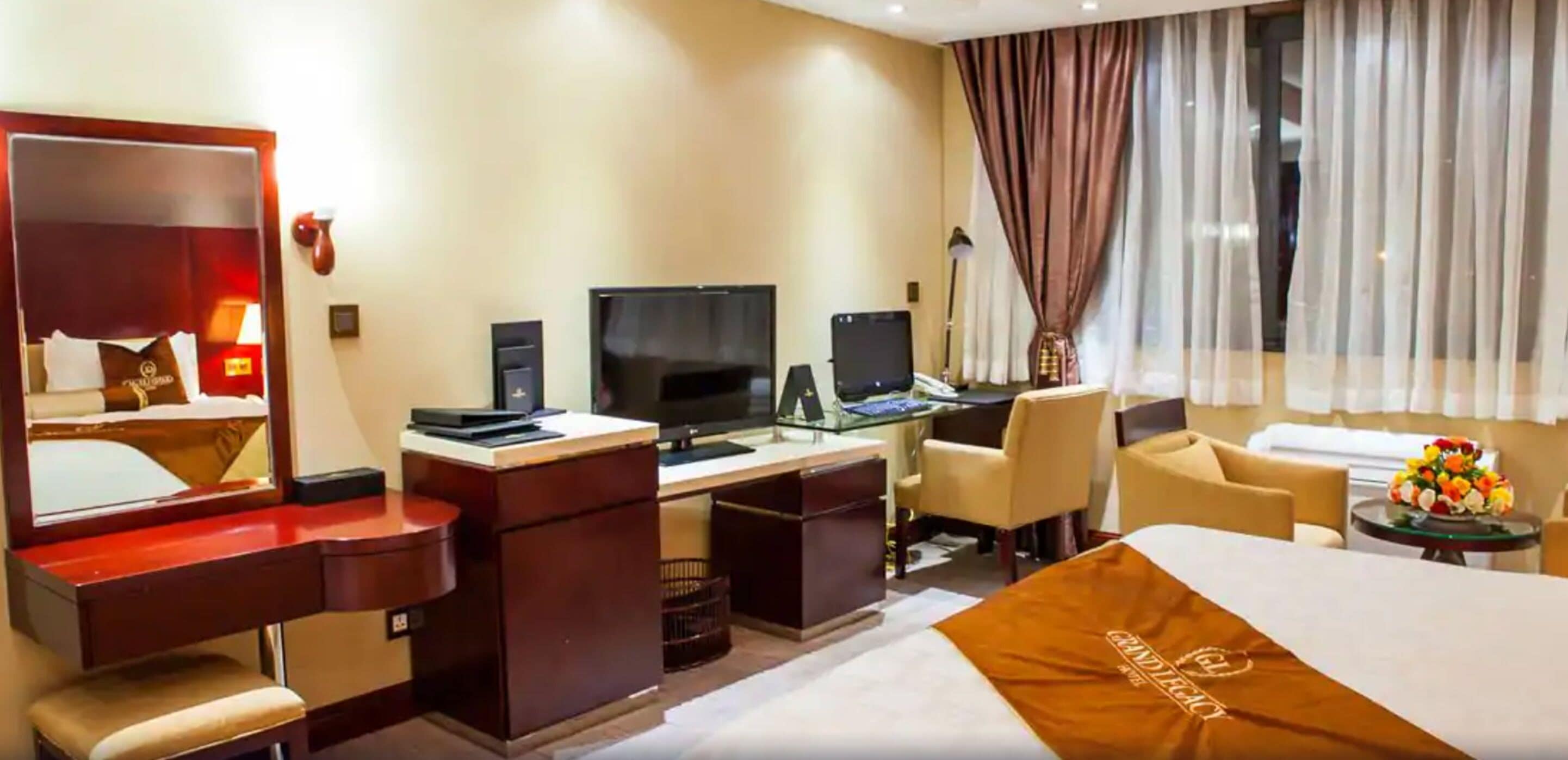 Habitación de hotel con una cama grande, un escritorio con ordenador, una silla, televisor y un espejo.