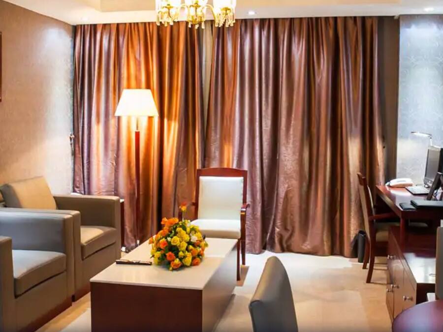 Habitación de hotel con sofá, sillones, un escritorio con computadora, una lámpara y un ventanal con cortinas.