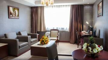 A modern hotel room with a sofa, a dining table with a flower arrangement, a TV, and a view of the city through large windows.