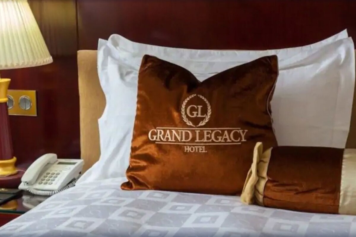 Una habitación de hotel con una cama, un almohadón con el logotipo de 'Grand Legacy Hotel', una lámpara de noche y un teléfono en la mesita.
