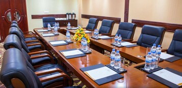 Un salón de conferencias con una larga mesa, sillas, botellas de agua y un arreglo floral.