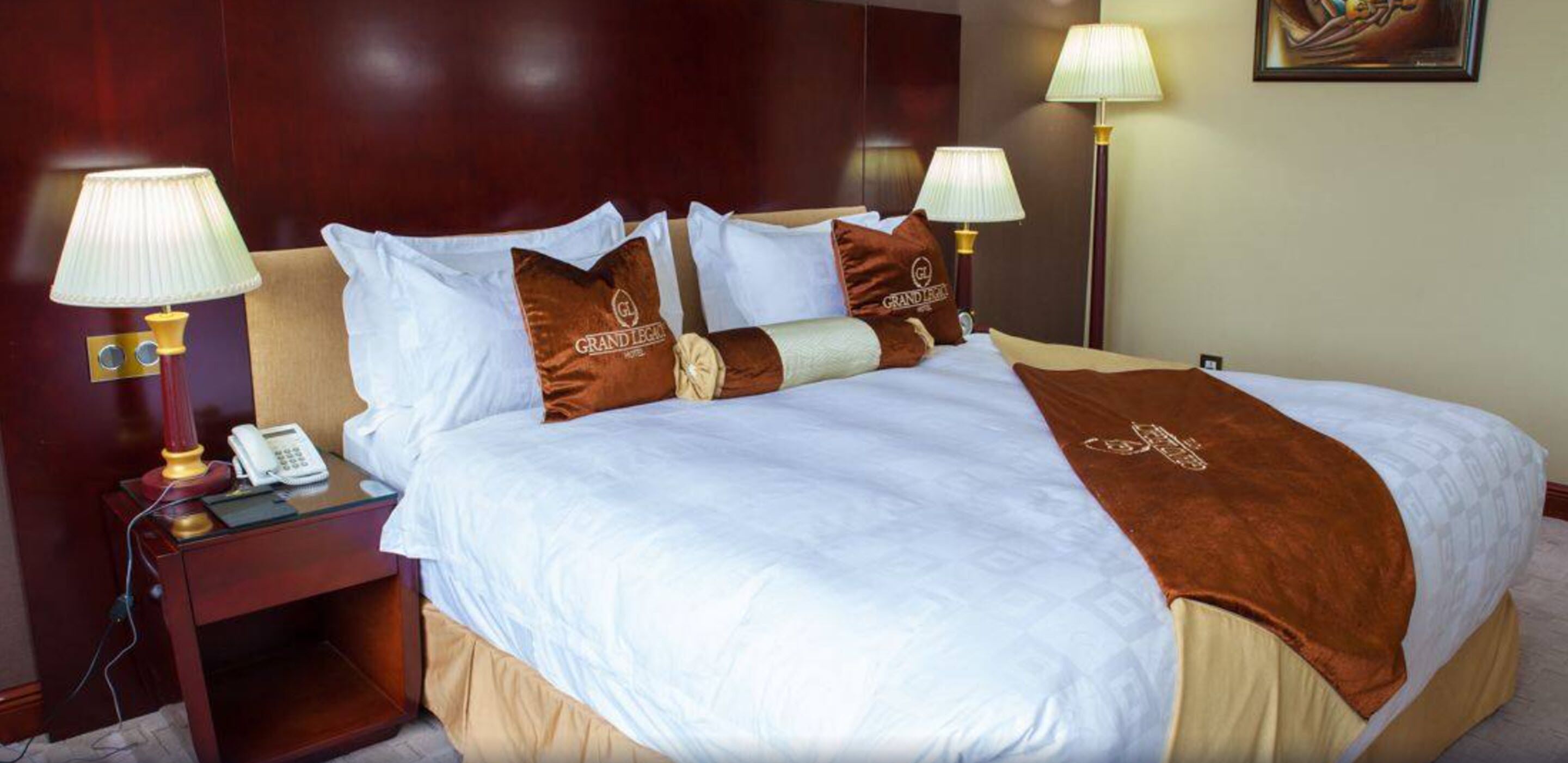 A hotel room with a neatly made bed, two bedside lamps, a nightstand with a phone, and a painting on the wall.