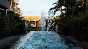 Una fuente en el centro de un estanque rodeado de vegetación y edificios al fondo.