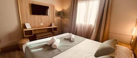 Una habitación de hotel con una cama, televisor montado en la pared, mesita de noche y ventana con cortinas.