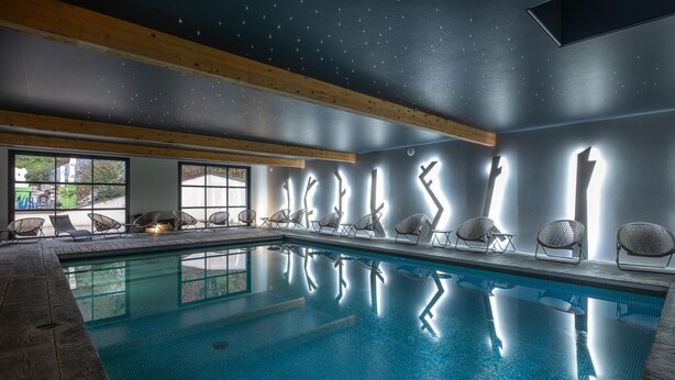 A swimming pool with chairs along the edge, wooden beams on the ceiling, and a view of the outdoors through large windows. - Le M Hôtel & Spa Honfleur (Honfleur)