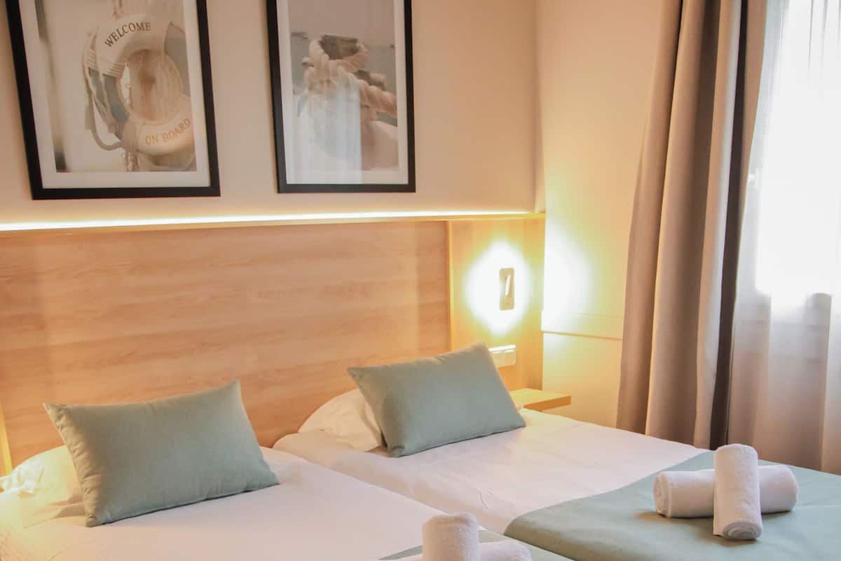 Ein Hotelzimmer mit zwei Betten, jeweils mit einem Kopfkissen und einem zusammengerollten Handtuch. An der Wand hängen gerahmte Bilder und auf dem Nachttisch steht eine Lampe.