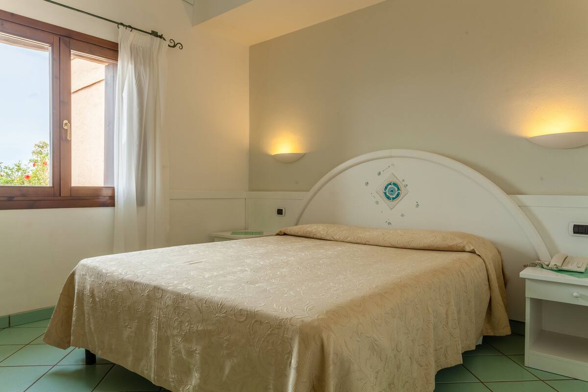 A bedroom with a bed, a nightstand, a wall-mounted light, and a window with a curtain.