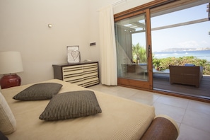 A bedroom with a bed, a dresser, a nightstand, and a view of the ocean through a sliding glass door. - Santo Stefano Resort & Spa (La Maddalena)