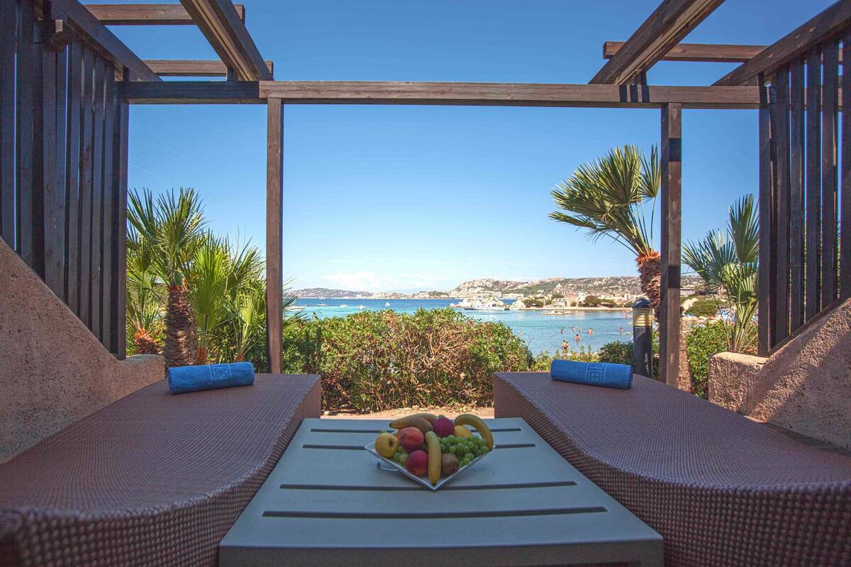 A balcony with wicker furniture, a table with a fruit bowl, and a scenic view of the sea and hills.