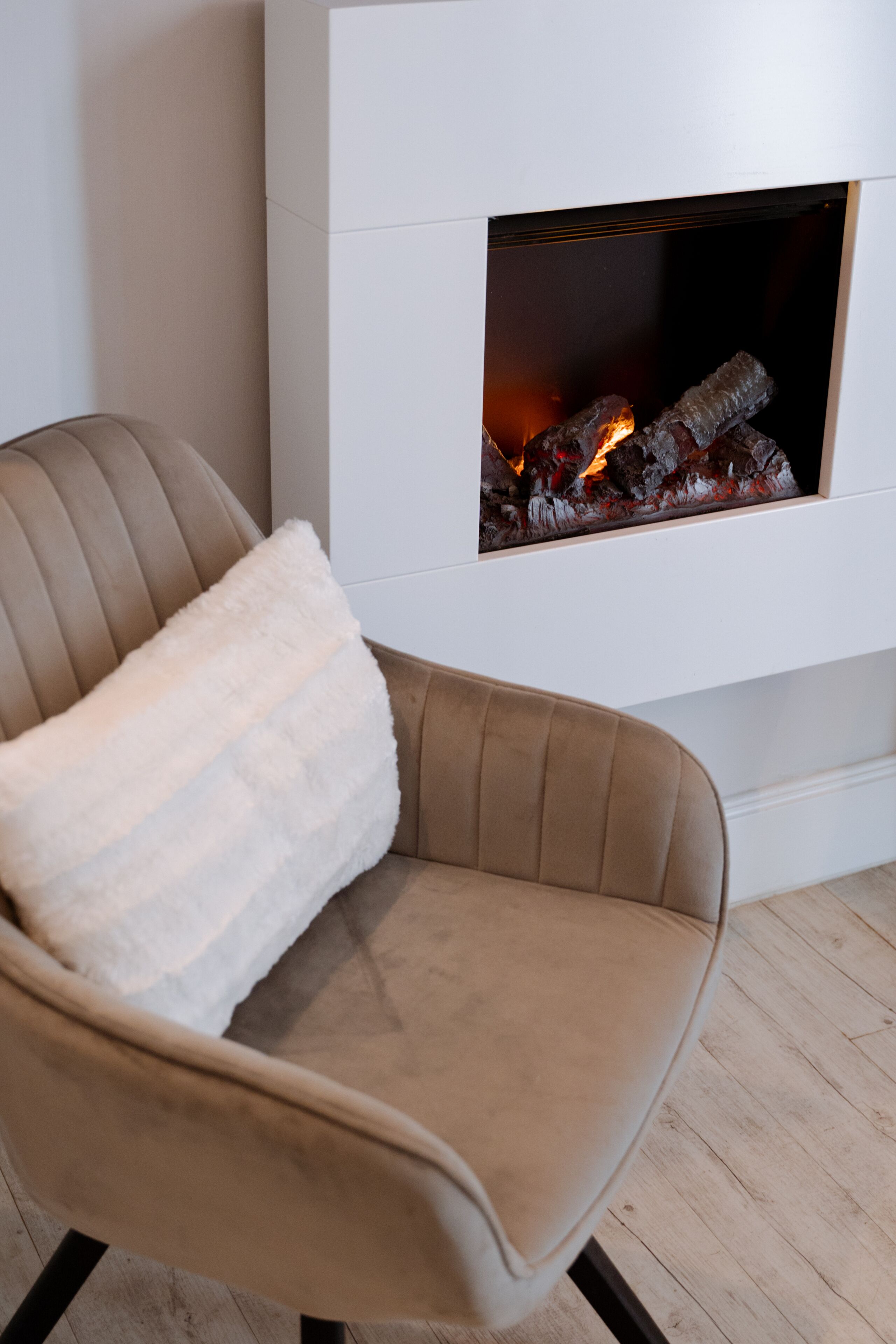 A modern living room with a beige armchair, a white fluffy pillow, and a white fireplace with logs.