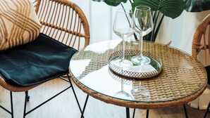 Una silla de mimbre con un cojín negro, una mesa de cristal con un plato decorativo y una planta de hojas grandes.
