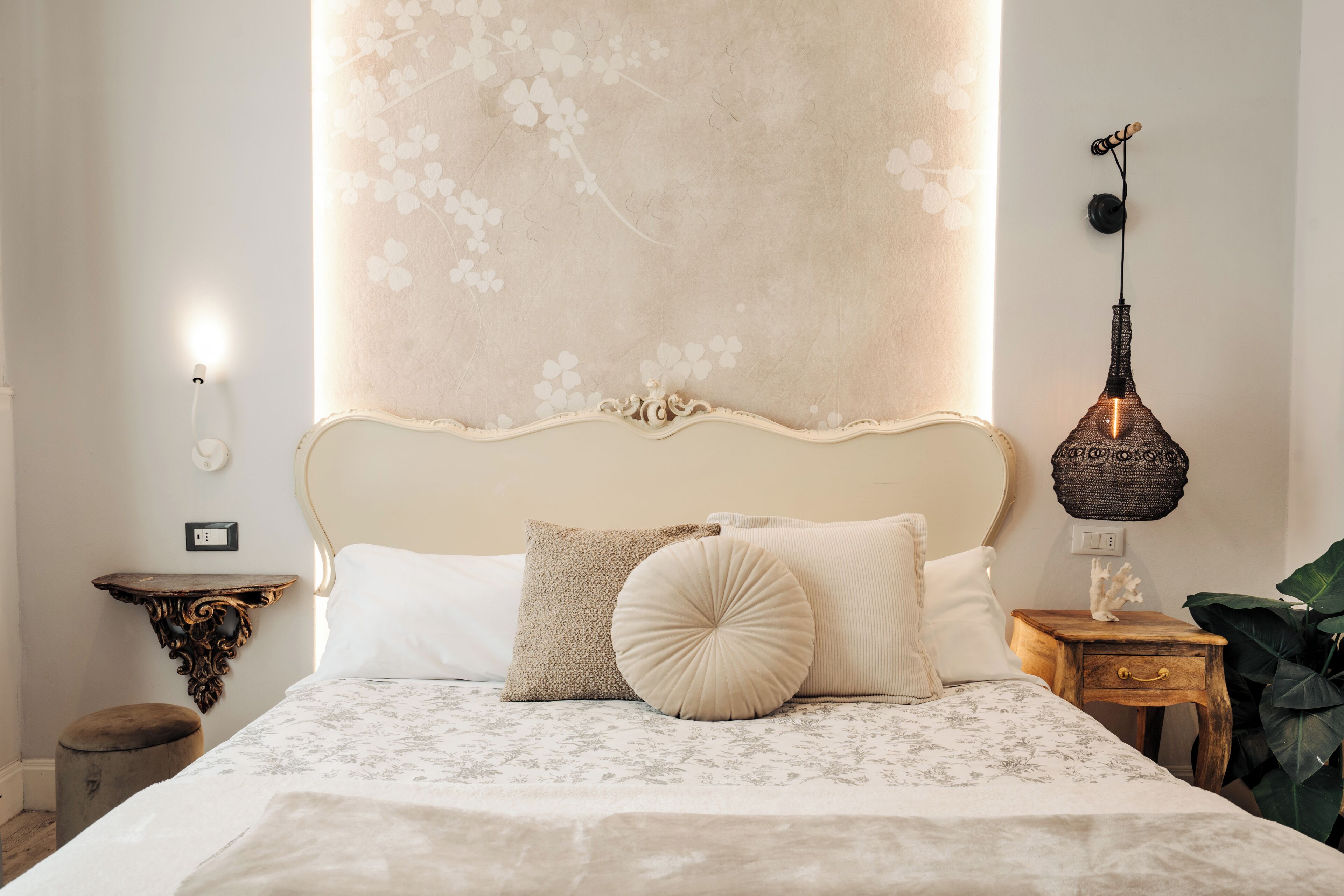 Una cama bien hecha con una colcha estampada con motivos florales, una pared decorativa con diseño floral, una lámpara de pared y una mesita de noche de madera con un cajón.