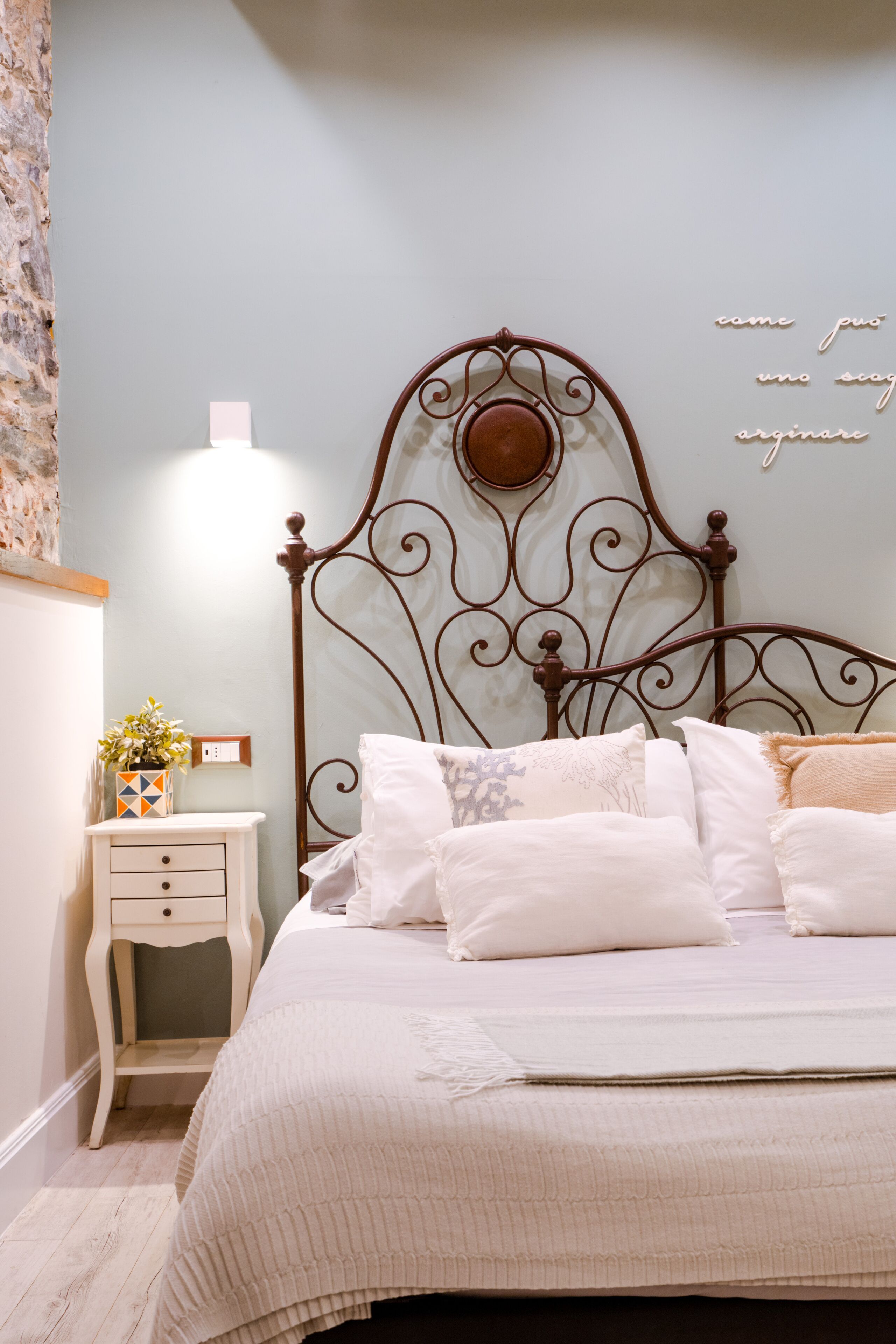 Un lit bien fait, avec une tête de lit en métal, une table de chevet ornée d’un vase de fleurs et une lampe fixée au mur.