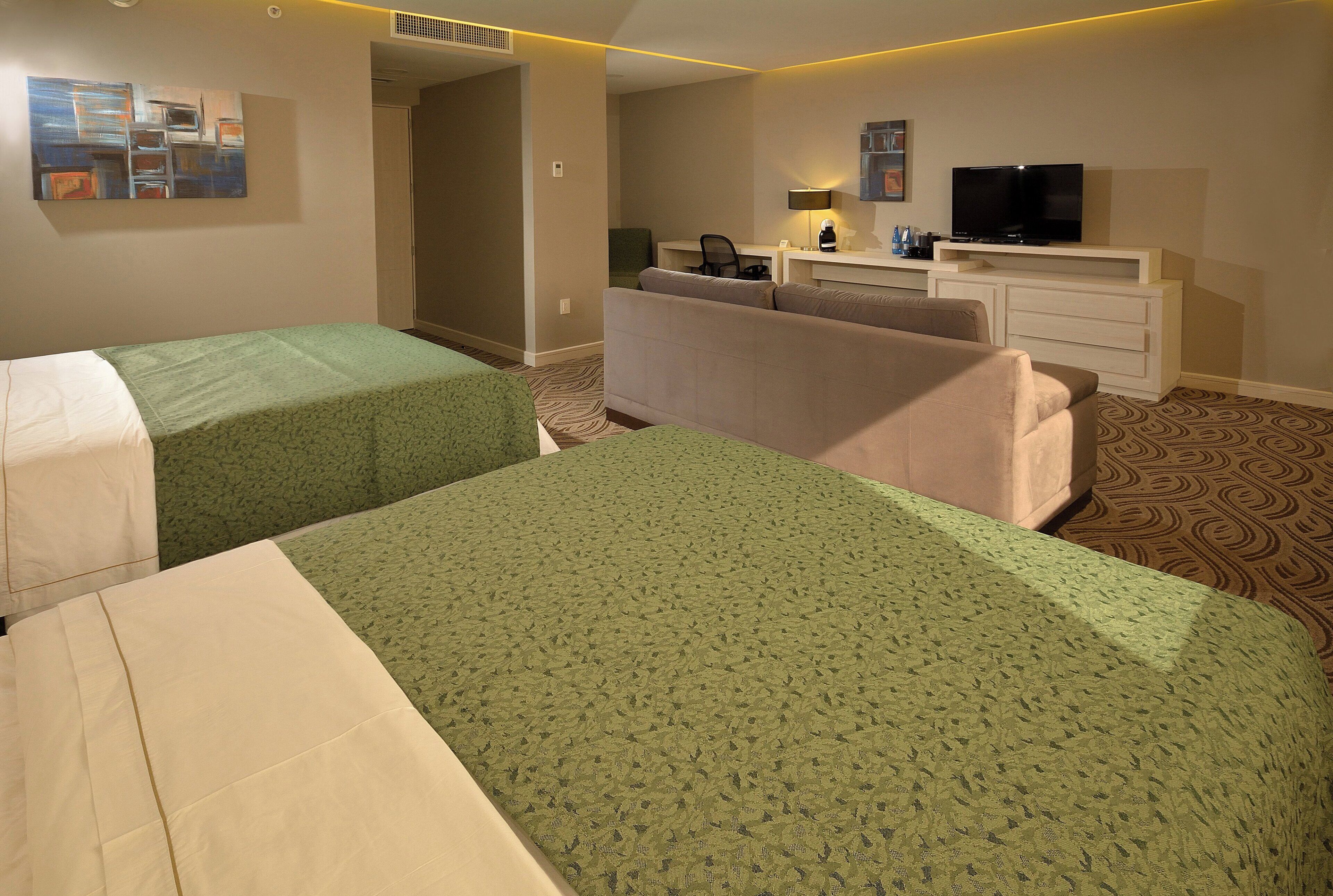 A hotel room with two single beds, a sofa, a desk with a computer, and a television.