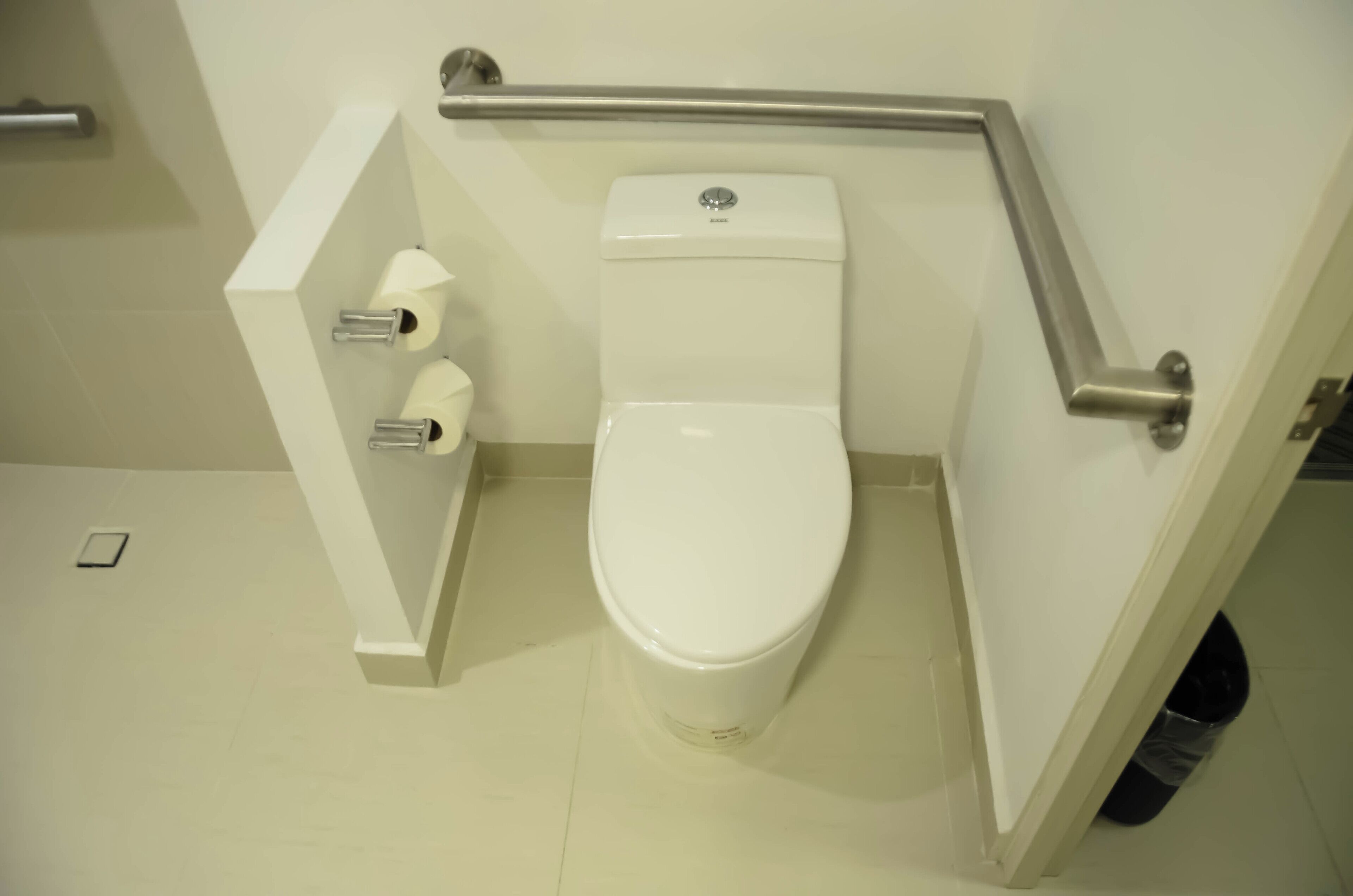 A white toilet with a handrail, a toilet paper holder, and a trash can.